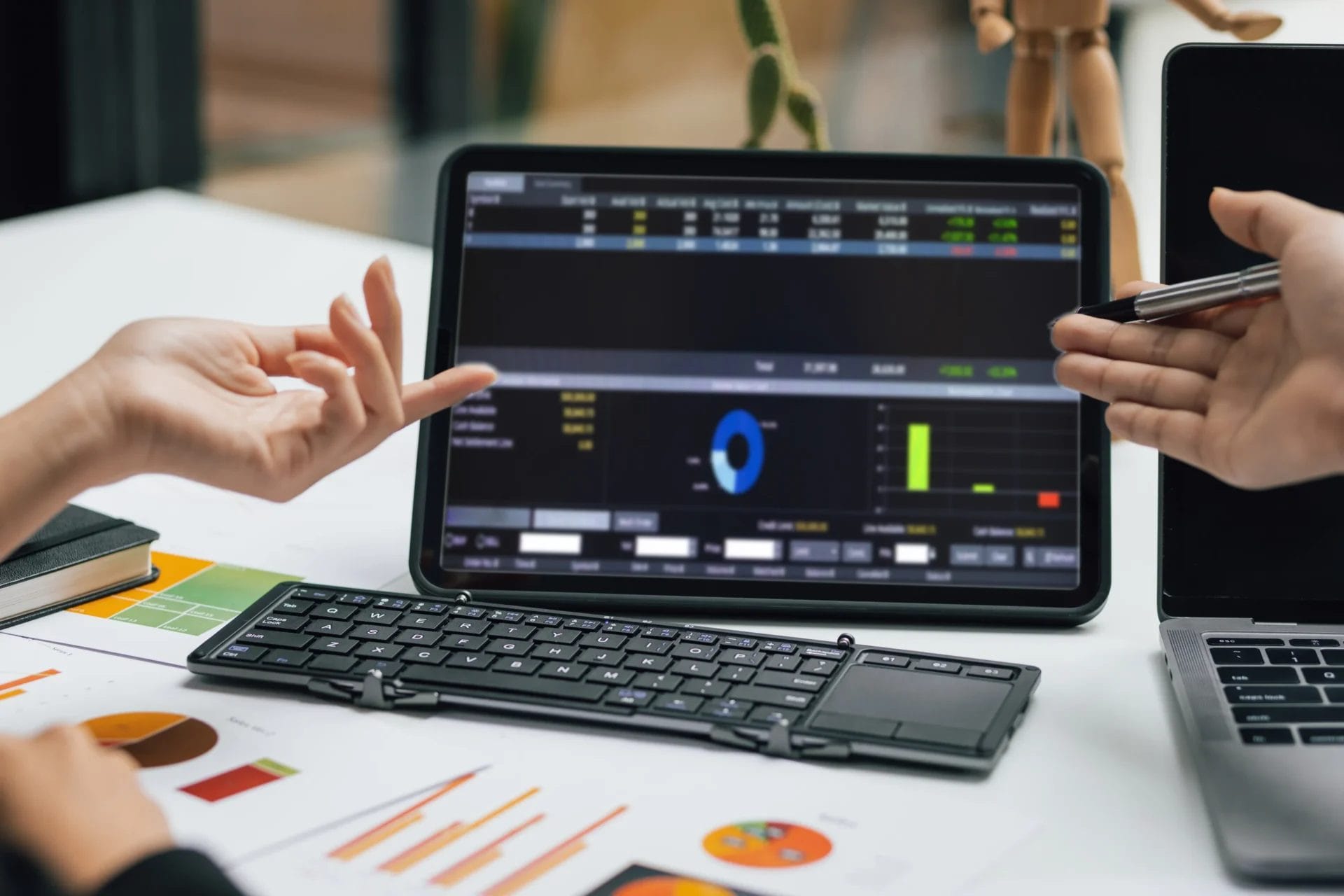 Tablet showing a lead tracker interface displaying business analytics and financial charts on a desk with a wireless keyboard, printed graphs, and two people gesturing at the data during a meeting. 