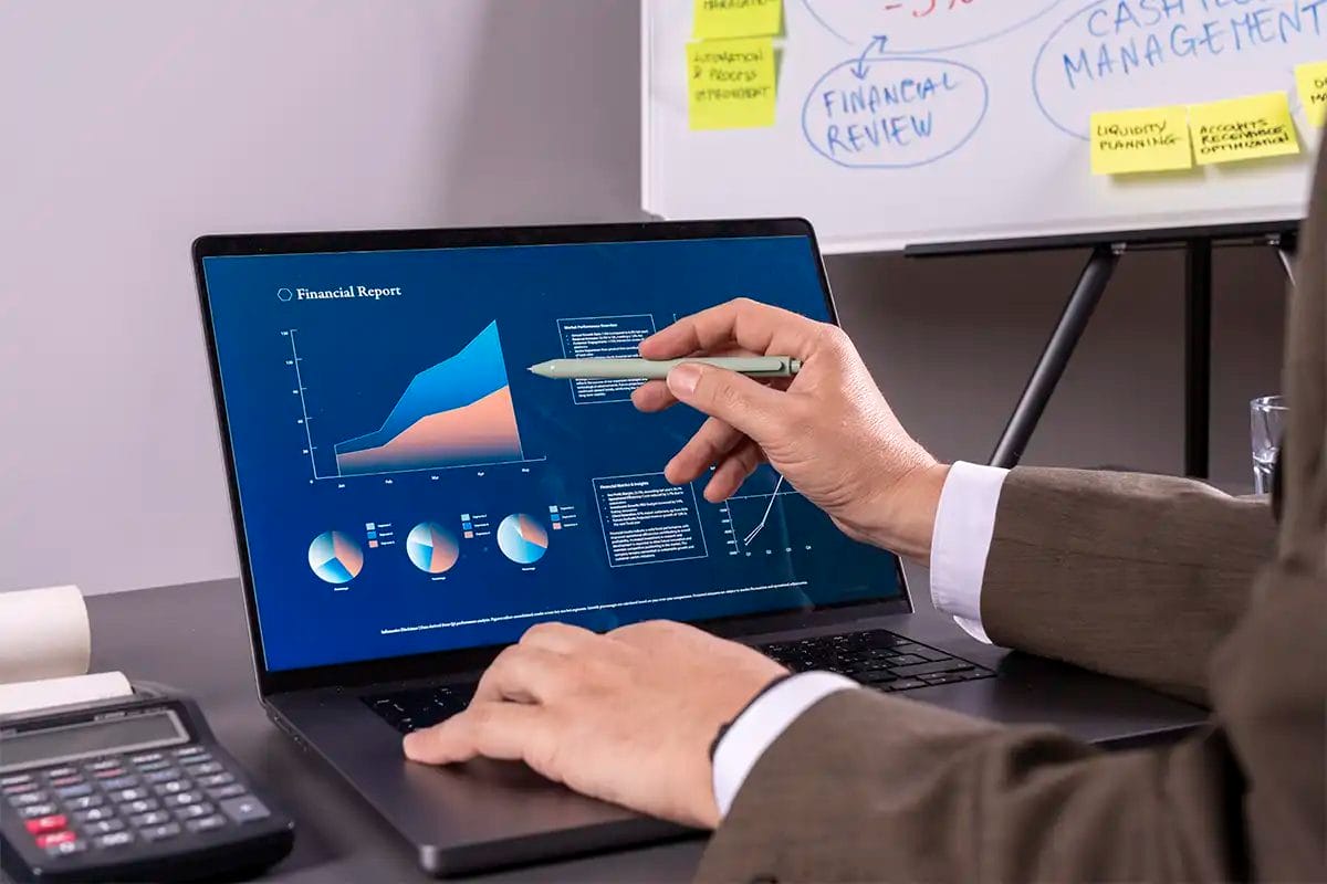 businessman pointing at a laptop screen displaying google ads services charts and reports, with a calculator and documents on the desk and a whiteboard filled with financial planning notes