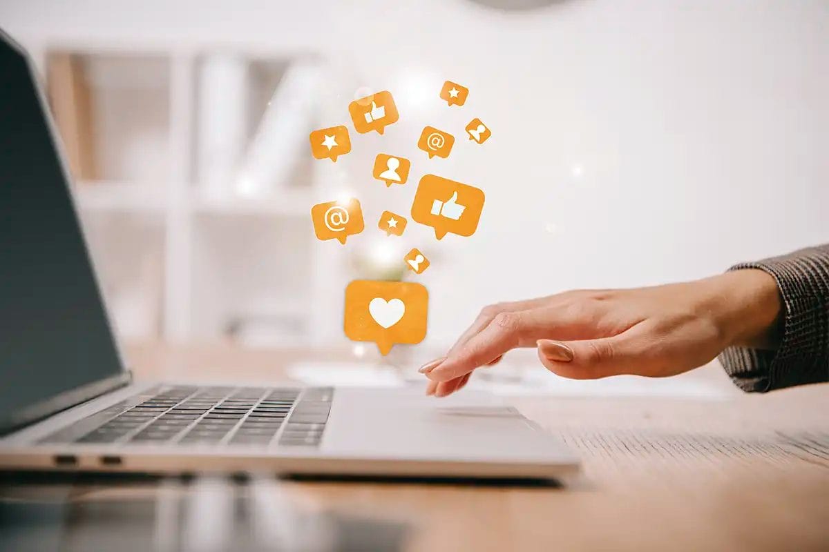 close up of a hand reaching toward a laptop keyboard with floating social media icons, including likes, hearts, mentions, and notifications, symbolizing social media advertising services