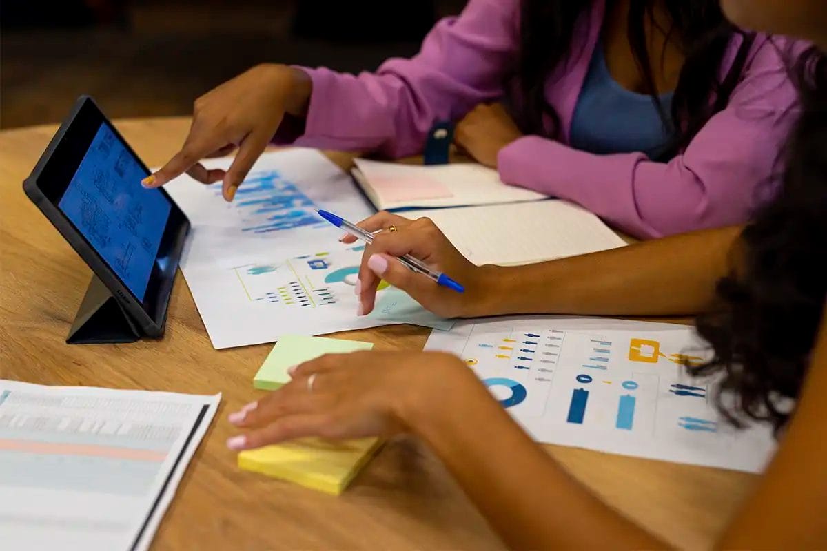group of people working together at a table, pointing at a tablet and reviewing printed charts and infographics with blue and yellow data visuals simbolizing off page seo services