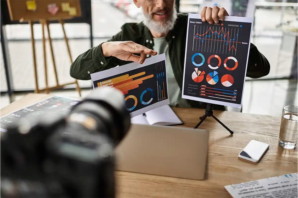 man presenting colorful printed charts and graphs on stands in front of a camera, with a laptop, notebook, and phone on the table, symbolizing a display advertising company presentation