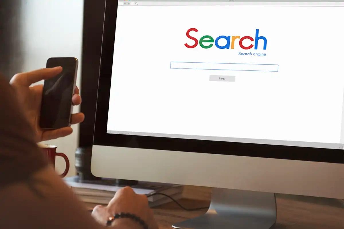 person holding a smartphone while using a desktop computer displaying a google homepage with a colorful ‘search’ logo, search bar, and enter button, suggesting online remarketing services