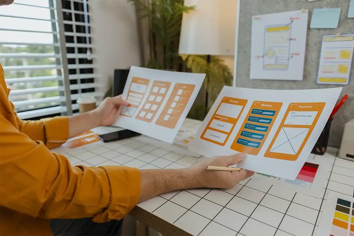 person reviewing mobile app wireframes and ui mockups on printed sheets at a design desk, surrounded by sketches, notes, and design tools, symbolizing a b testing in google ad services