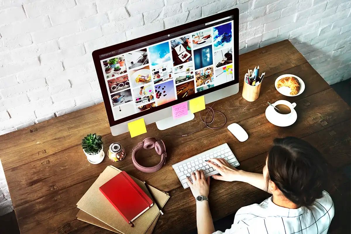 person working on a desktop computer displaying a photo gallery, surrounded by notebooks, headphones, coffee, and a croissant on a social media marketing agency las vegas