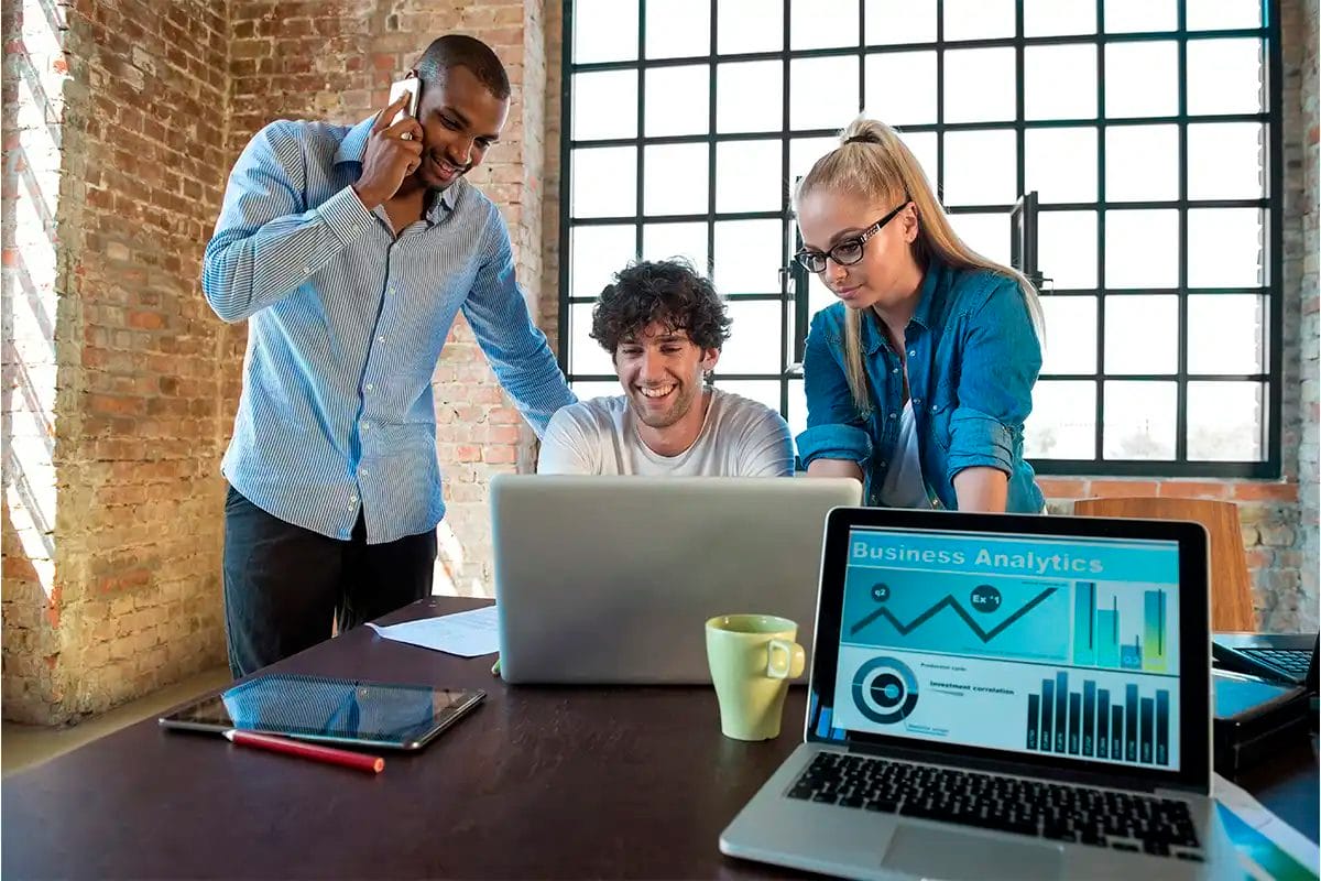 three young professionals around a laptop in a modern office, with another laptop in the foreground displaying a remarketing services's analytics dashboard with charts and graphs