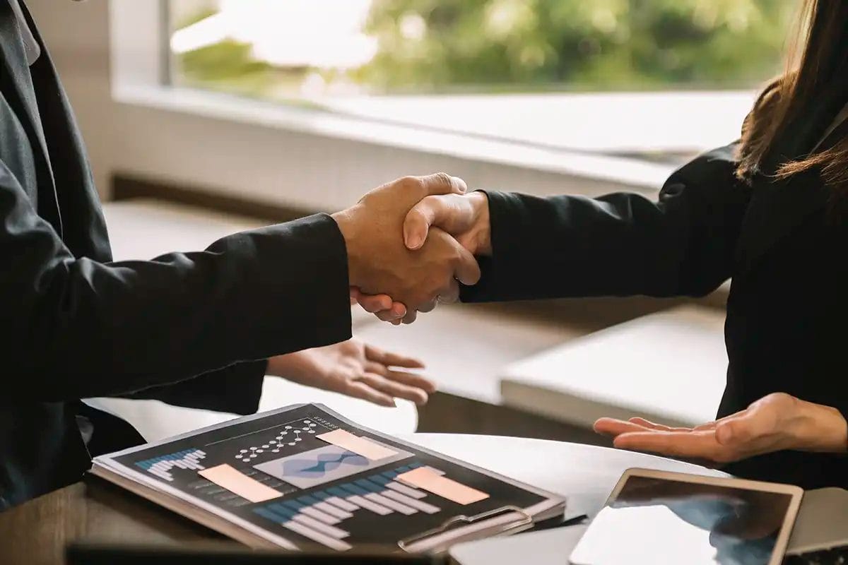 two business professionals in dark suits shaking hands across a table with business charts, documents, and a tablet, symbolizing a successful business partnership for remarketing services