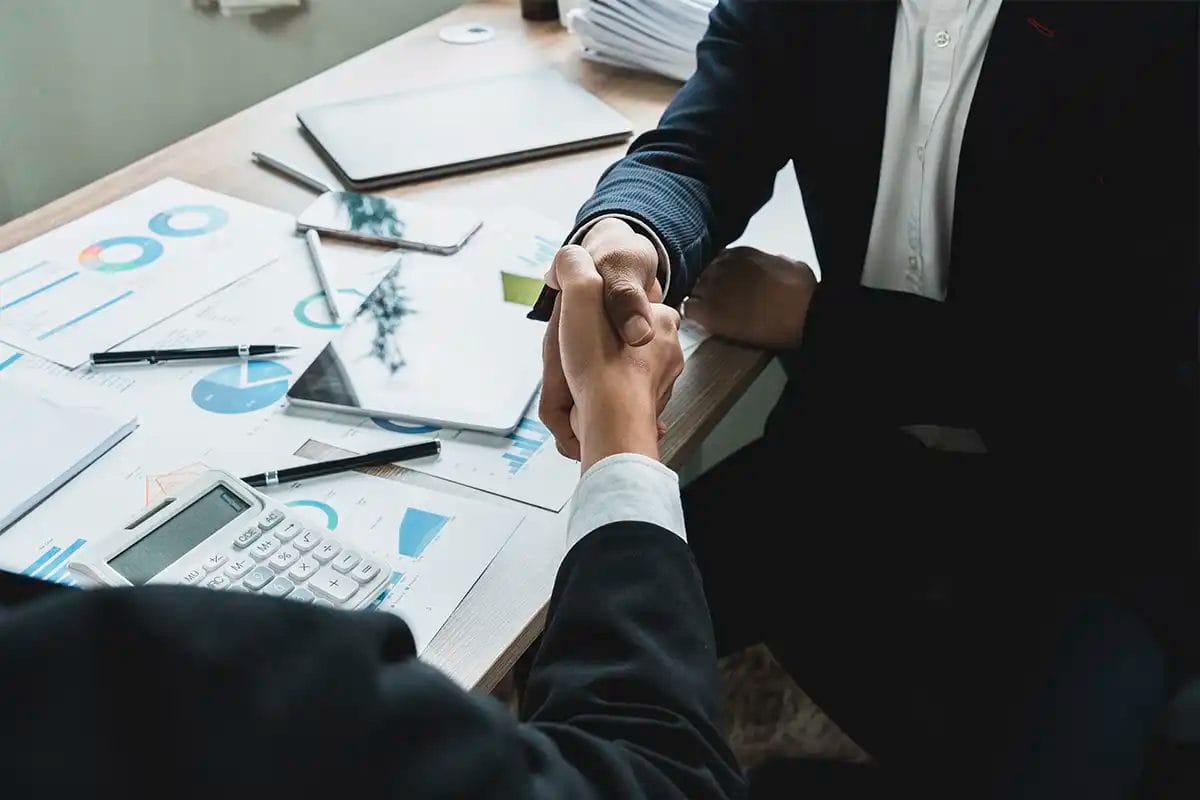 two business professionals shaking hands across a desk covered with financial documents, charts, and devices, symbolizing a business agreement with social media advertising services