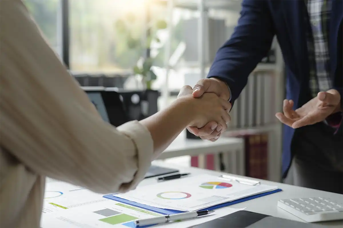 two business professionals shaking hands over a table with charts and reports, symbolizing a successful agreement with a ada compliance agency, modern office setting