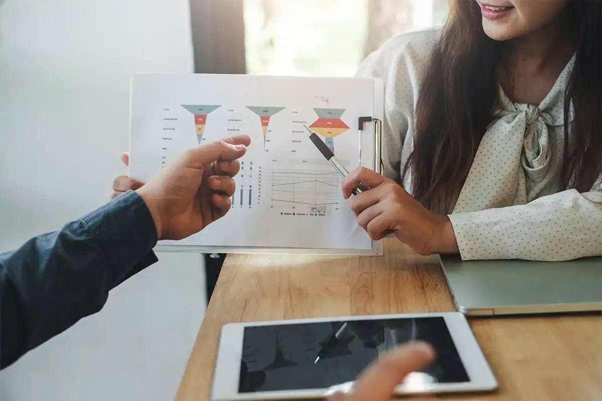 two colleagues discussing a printed sales funnel and data charts on a clipboard during a meeting, with a tablet and laptop on the table, symbolizing lead tracking in google ads services