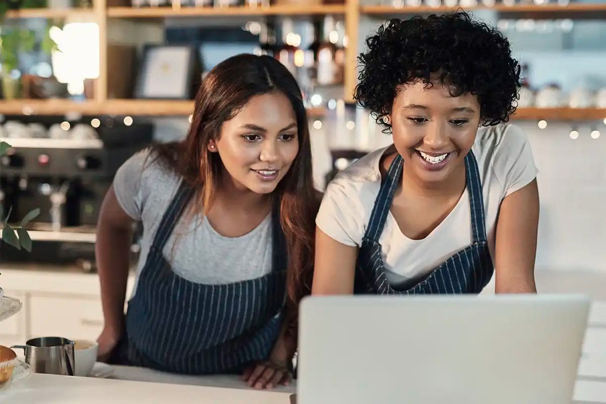 two smiling café workers wearing aprons, looking at a laptop screen, indicating the work of a google maps marketing agency