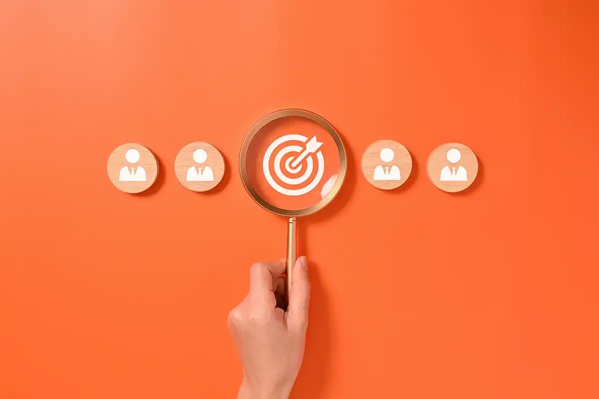 wooden blocks on an orange background with a central bullseye target surrounded by icons of a handshake, bar chart, computer graph, gears, clipboard, and lightbulb, symbolizing display adversing