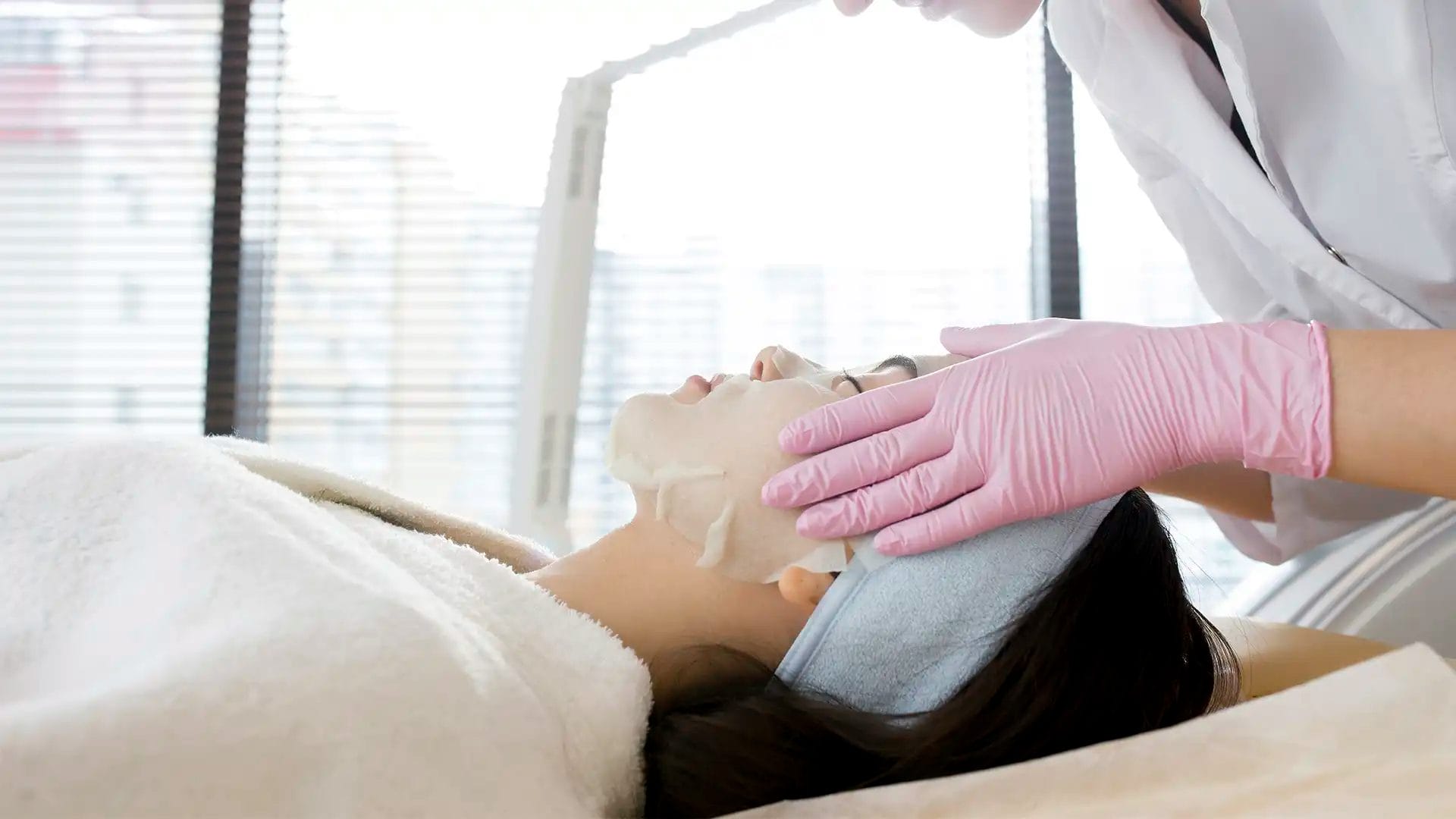 aesthetician wearing pink gloves applying a sheet mask to a client’s face during a facial treatment at a bright medical spa digital marketing agency