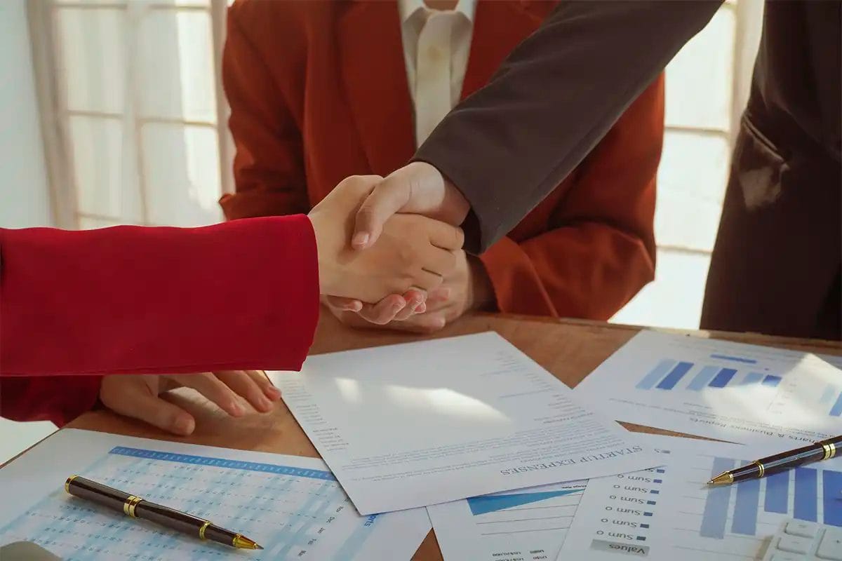 two business professionals shaking hands over a desk covered with documents, bar charts, and pens a third person is seated in the background agreement with a google business profile agency