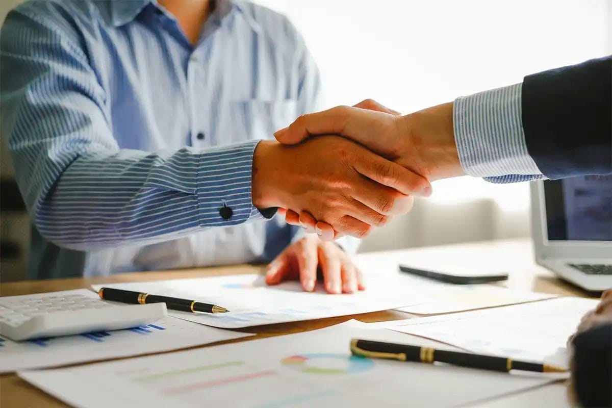 two professionals in striped business shirts shaking hands over a desk covered with financial documents, pens, a calculator, and a laptop simbolizing agreement with a google maps agency