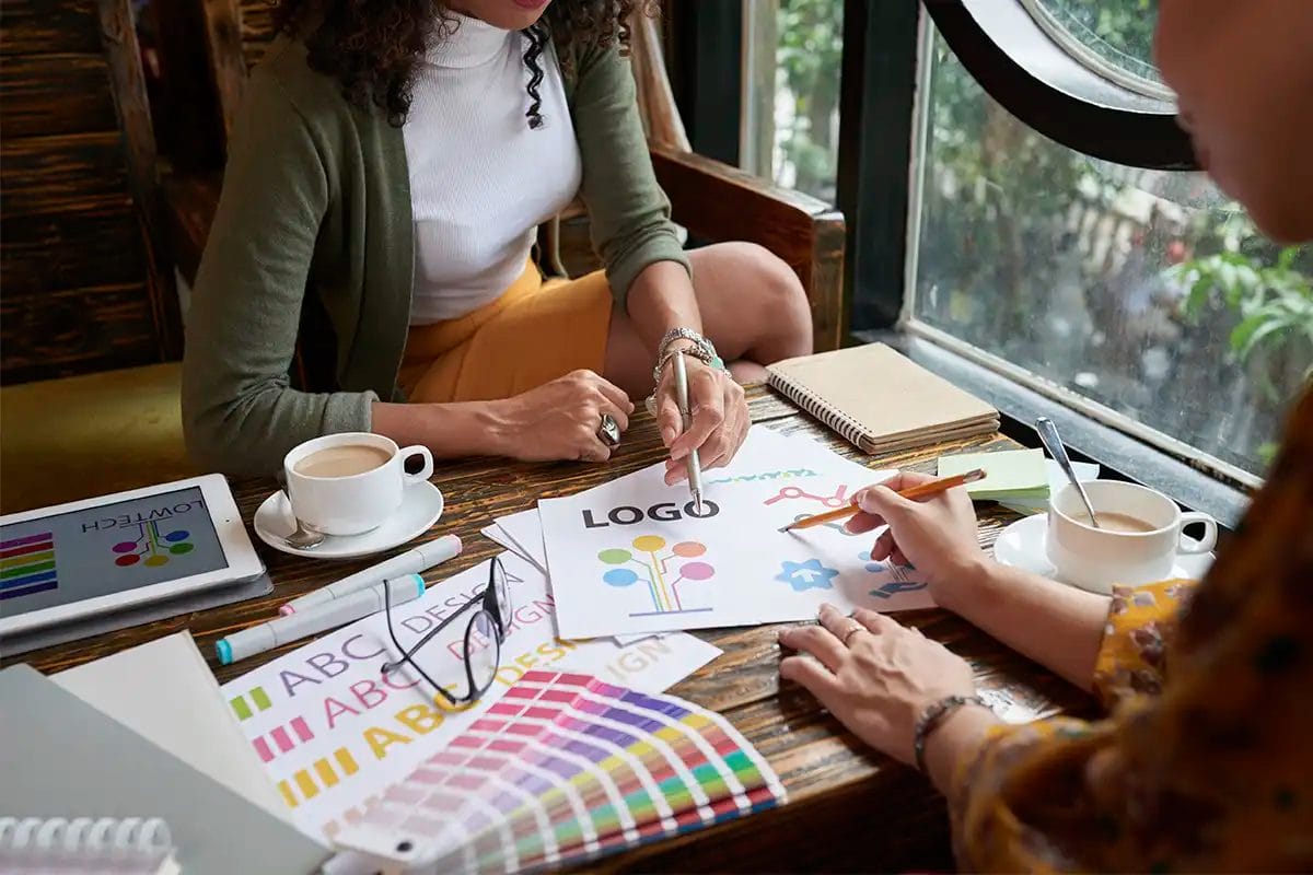 two women collaborating on a logo design at a rustic wooden table by a large window referring to the work of social media content creation services