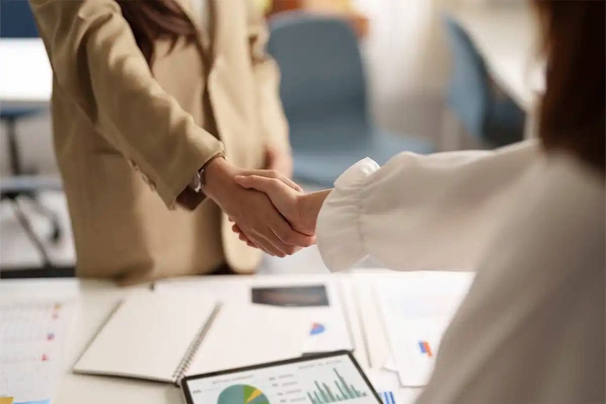 two women in business attire shaking hands in a modern office setting a desk in the foreground is covered with a tablet displaying charts, a notebook, and financial documents simbolizing agreement with a Google My Business Post service.